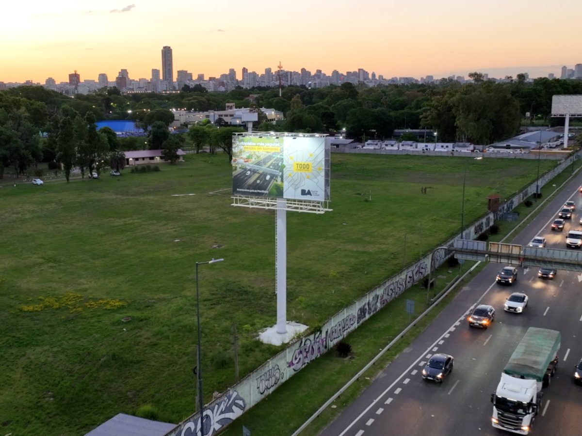 Vista aérea de un cartel del Gobierno de la Ciudad con la pieza «Autopistas más seguras con peajes sin barreras» junto a una autopista, al atardecer